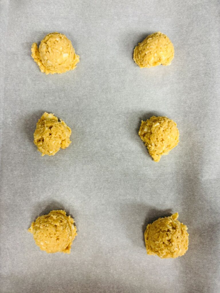 Six evenly spaced scoops of unbaked cookie dough are placed on a sheet of parchment paper, ready to be baked.