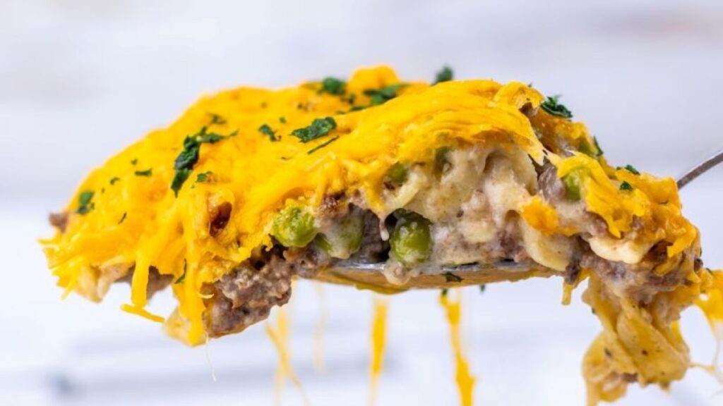 A close-up of a spoonful of Amish country casserole topped with melted cheddar cheese, showing ground meat, green peas, and creamy mashed potatoes, garnished with chopped parsley.