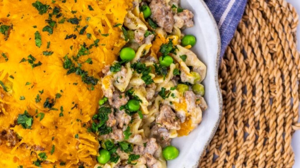 A plate of casserole with a golden, shredded topping on one side and a creamy mix of pasta, peas, and ground meat on the other, garnished with chopped parsley, on a woven placemat.