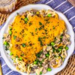 A plate of Amish country casserole topped with shredded cheddar cheese and chopped parsley, served with ground meat, peas, and a creamy sauce, on a striped cloth beside a gold fork and a glass of iced drink.