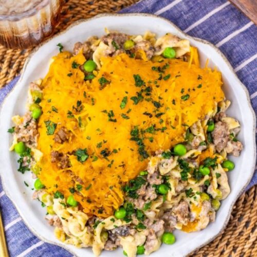 A plate of Amish country casserole topped with shredded cheddar cheese and chopped parsley, served with ground meat, peas, and a creamy sauce, on a striped cloth beside a gold fork and a glass of iced drink.
