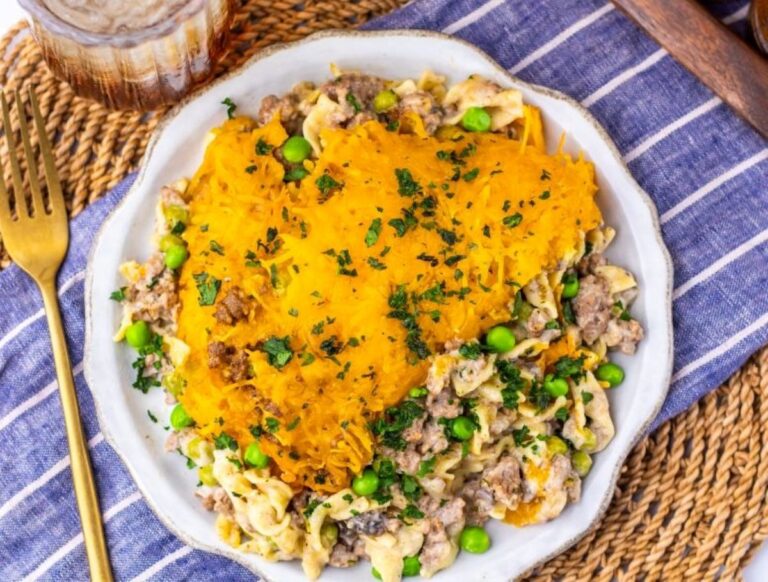 A plate of Amish country casserole topped with shredded cheddar cheese and chopped parsley, served with ground meat, peas, and a creamy sauce, on a striped cloth beside a gold fork and a glass of iced drink.