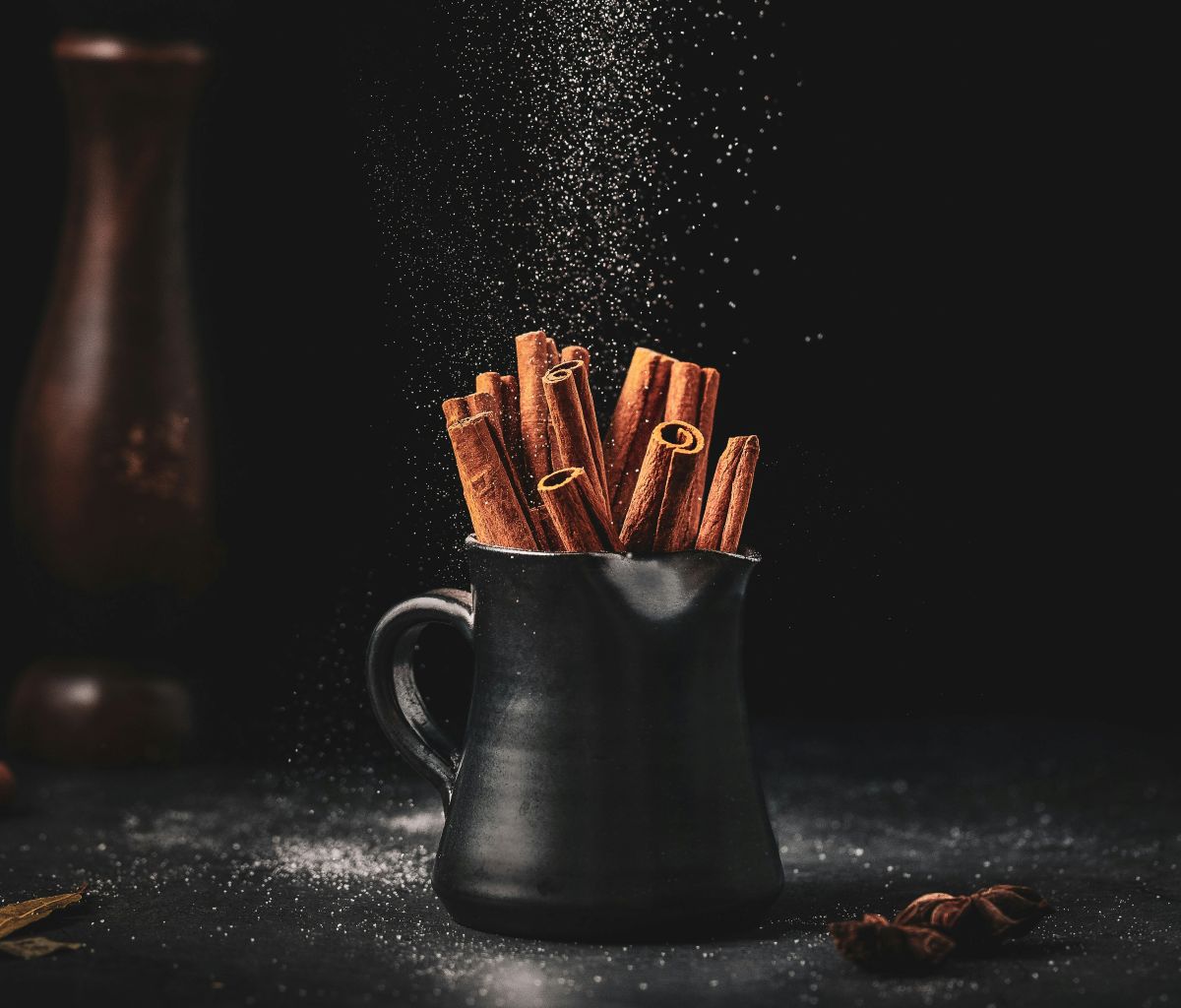 A black ceramic mug filled with cinnamon sticks sits on a dark surface, surrounded by scattered spices and powder. A gentle sprinkle of powder falls from above, creating a dramatic, moody atmosphere.