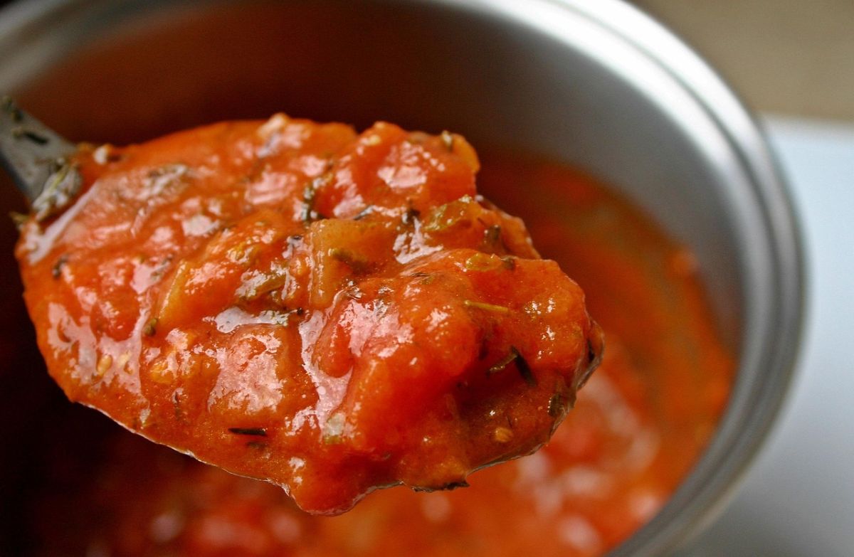 A close-up of a spoonful of thick, chunky tomato sauce with herbs, held above a pot filled with the same sauce.