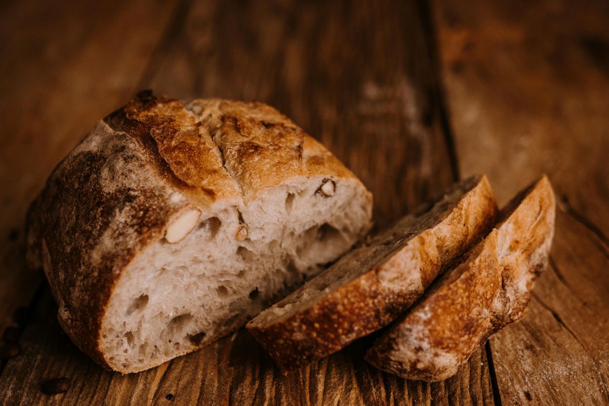 A loaf of rustic artisan bread with a golden crust sits on a wooden surface. Two slices have been cut from the loaf, revealing a soft, airy interior with some visible nuts.