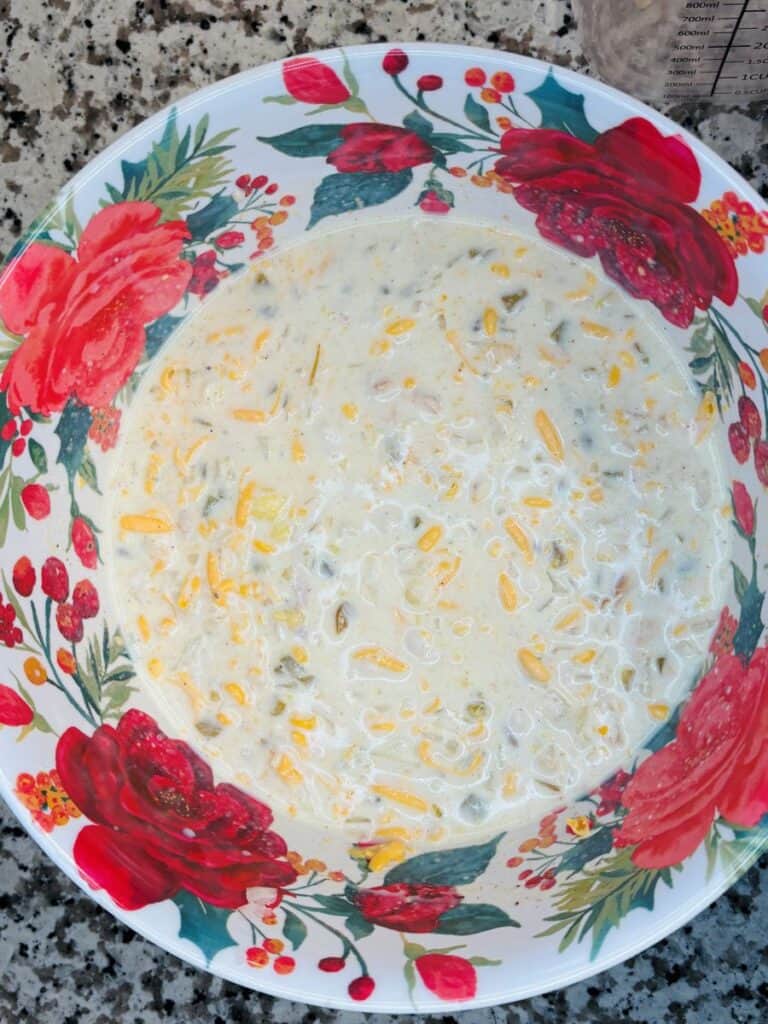 A festive floral bowl on a granite countertop is filled with a creamy mixture containing shredded cheese and chopped green chiles.