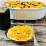 A plate of creamy casserole topped with melted cheddar cheese and black olive slices sits beside a jar of dark soda. A larger serving dish of the same casserole is in the background on a wooden table. A fork rests on the plate.