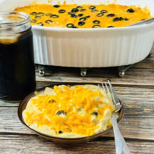 A plate of creamy casserole topped with melted cheddar cheese and black olive slices sits beside a jar of dark soda. A larger serving dish of the same casserole is in the background on a wooden table. A fork rests on the plate.