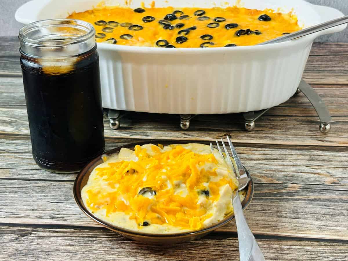 A plate of creamy casserole topped with melted cheddar cheese and black olive slices sits beside a jar of dark soda. A larger serving dish of the same casserole is in the background on a wooden table. A fork rests on the plate.