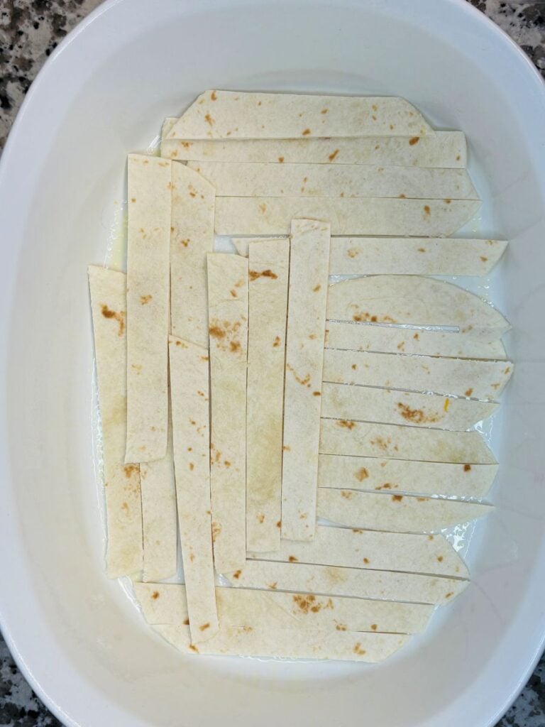 Strips of flour tortilla are arranged in a single layer in the bottom of a white casserole dish, ready for baking. The dish is on a granite countertop.