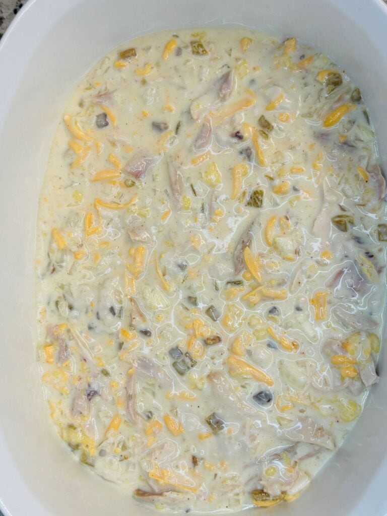 A close-up of a baking dish filled with an unbaked mixture of shredded chicken, yellow cheese, chopped green chilies, and a creamy white sauce.