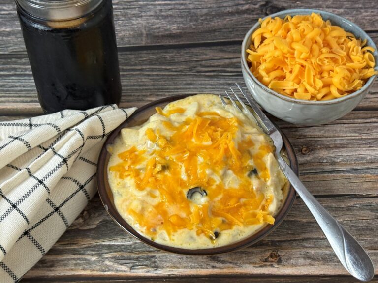 A plate of cheesy casserole topped with melted cheddar, next to a bowl of shredded cheese, a jar of dark beverage, and a checkered cloth on a wooden table. A fork rests on the plate.