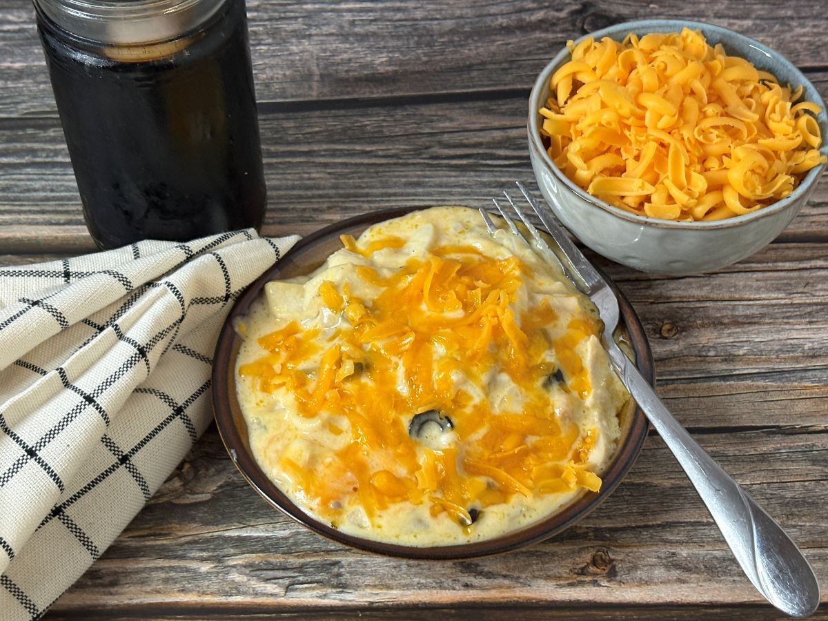 A plate of cheesy casserole topped with melted cheddar, next to a bowl of shredded cheese, a jar of dark beverage, and a checkered cloth on a wooden table. A fork rests on the plate.
