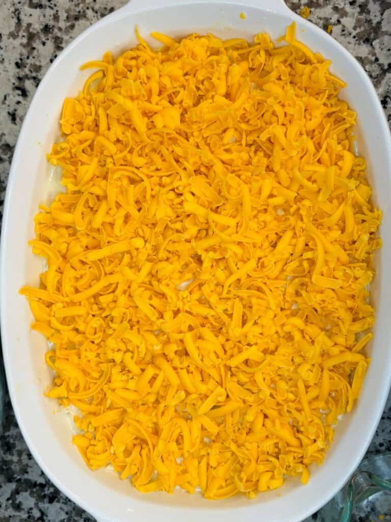 A white oval baking dish filled with an even layer of shredded yellow cheddar cheese, sitting on a speckled granite countertop.
