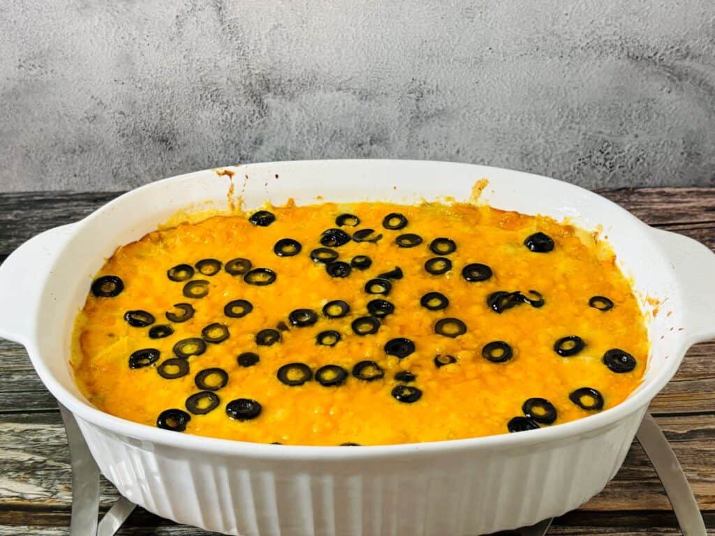 A white casserole dish filled with a baked dish topped with melted orange cheese and sliced black olives, placed on a rustic wooden surface with a gray textured background.