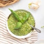 A bowl of green pesto garnished with fresh basil leaves, with a spoon in the bowl, sits on a striped cloth. Fresh basil, garlic, and a woven placemat are nearby on a white surface.