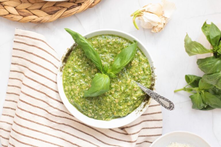 A bowl of green pesto garnished with fresh basil leaves, with a spoon in the bowl, sits on a striped cloth. Fresh basil, garlic, and a woven placemat are nearby on a white surface.