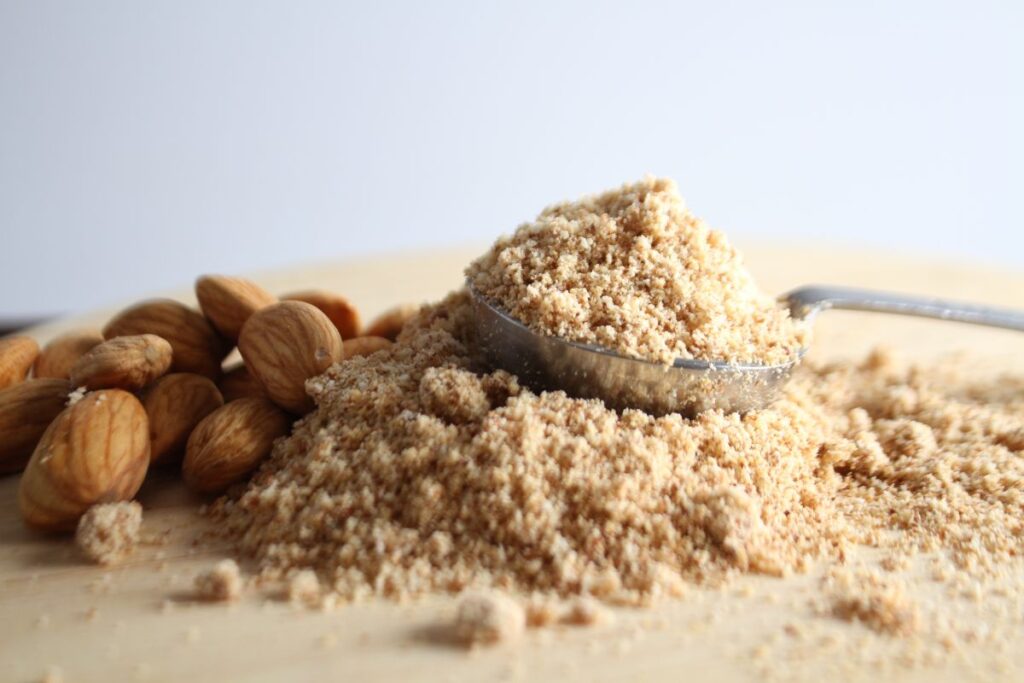 A spoonful of almond flour rests on a wooden surface, with a small pile of the flour spilling over and several whole almonds scattered nearby.