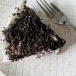A slice of cake topped with crushed chocolate cookies sits on a patterned white plate next to a silver fork.