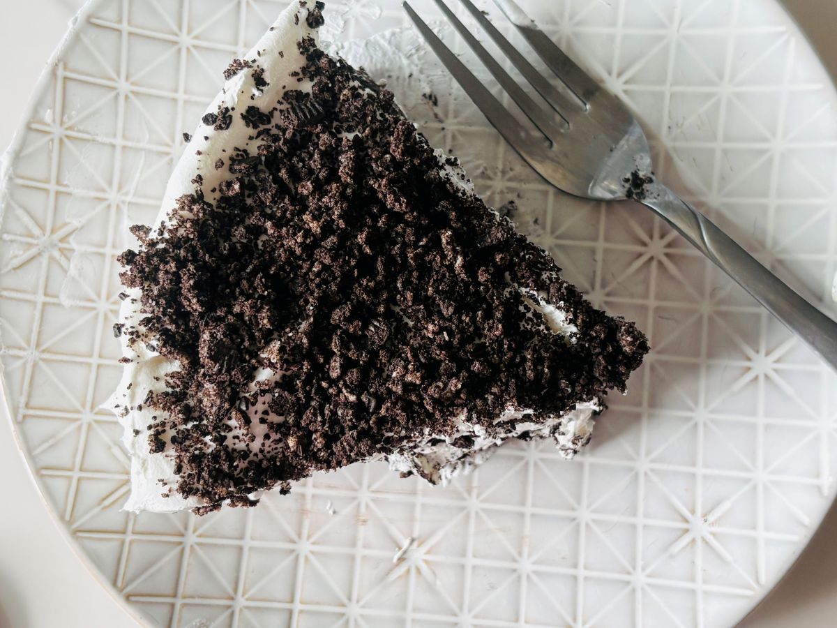 A slice of cake topped with crushed chocolate cookies sits on a patterned white plate next to a silver fork.