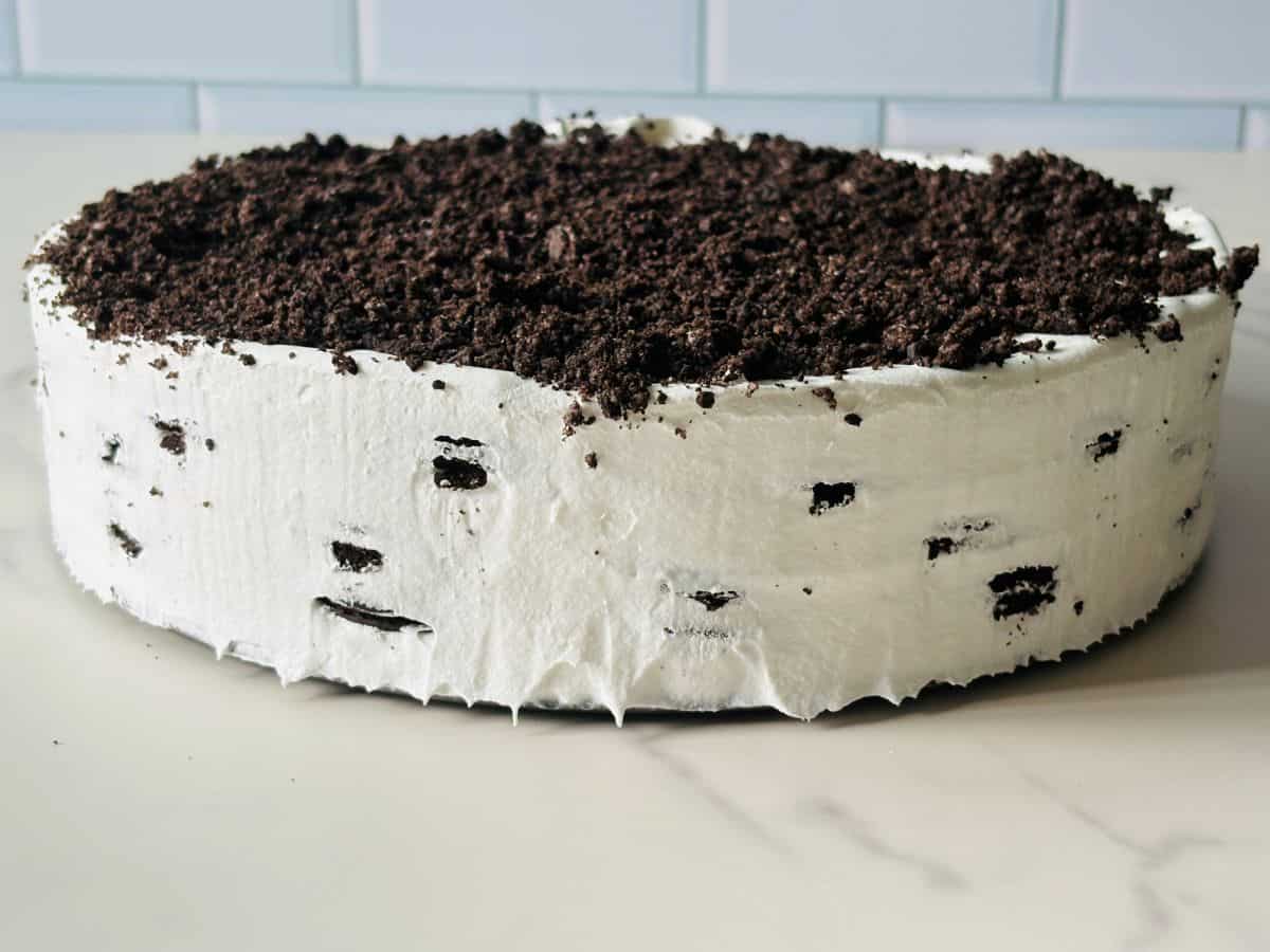 A round cake with white frosting and crushed chocolate cookies on top, set on a light-colored surface in front of a tiled wall.