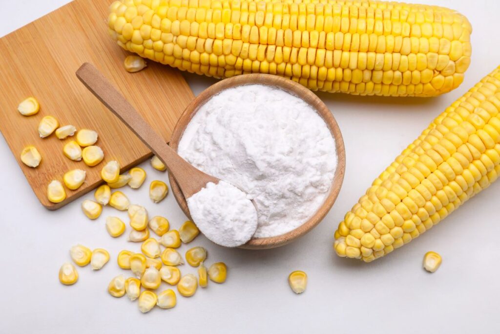 Two fresh corn cobs, loose corn kernels, and a bowl of white cornstarch with a wooden spoon are arranged on a wooden board and white surface.