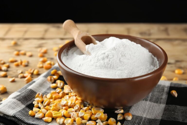A brown bowl filled with white corn starch powder, with a wooden scoop resting inside. Yellow corn kernels are scattered around the bowl on a black-and-white checkered cloth and wooden surface.