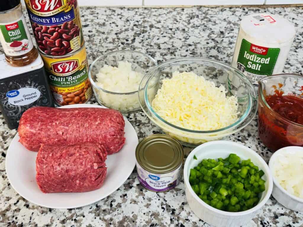 Ingredients for bean dip arranged on a countertop, including ground beef, canned beans, shredded cheese, chopped onions, green bell pepper, tomato sauce, black olives, chili powder, pepper, and salt.