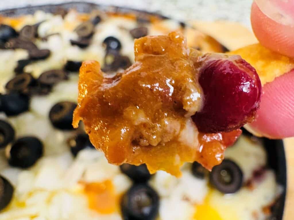 A close-up of a hand holding a chip topped with chili and a cherry, with a dish of chili, melted cheese, and sliced black olives in the background.