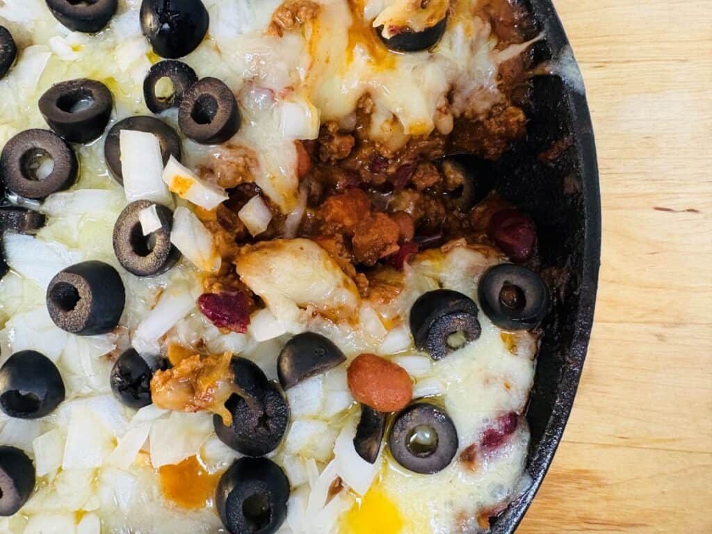 A close-up of a skillet dish topped with melted cheese, chopped onions, sliced black olives, beans, and ground meat on a wooden surface. Some of the dish has been scooped out.