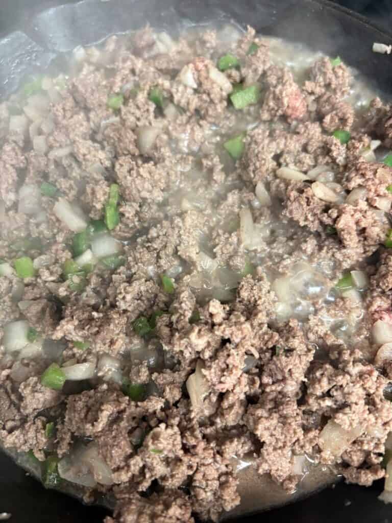 Ground beef cooking in a pan with diced onions and green peppers, with steam rising from the mixture.