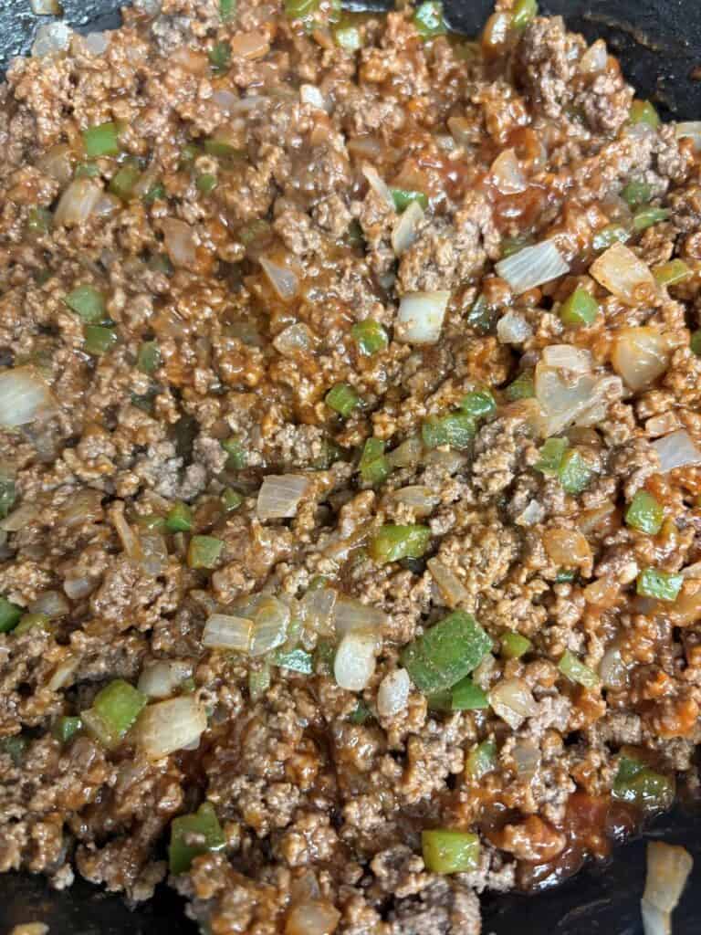 Ground beef cooking in a pan with diced onions, green bell peppers, and a reddish sauce, creating a savory mixture with visible cooked meat and vegetable pieces.