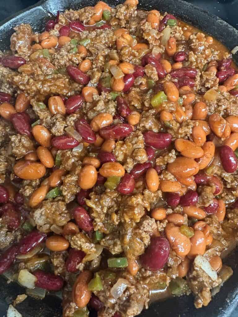 A close-up of a skillet filled with chili, featuring ground meat, kidney beans, pinto beans, chopped onions, and green peppers in a thick, spicy sauce.