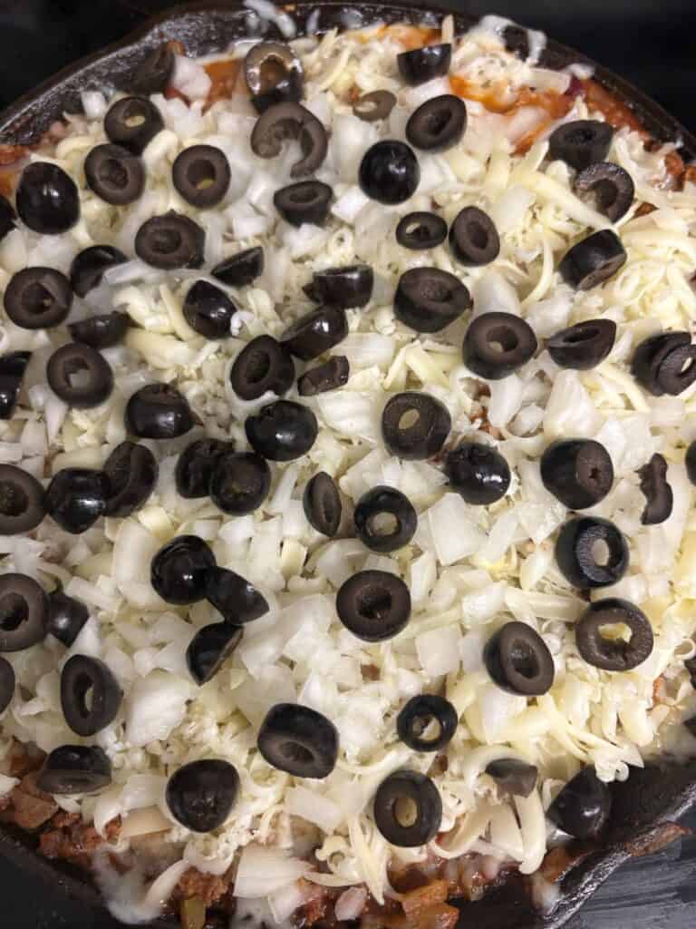 A close-up of an unbaked pizza topped with chopped onions, shredded mozzarella cheese, and sliced black olives. The ingredients are spread evenly over a tomato sauce layer on a thick crust.