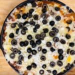A skillet dip topped with melted cheese, chopped onions, and sliced black olives on a wooden surface.