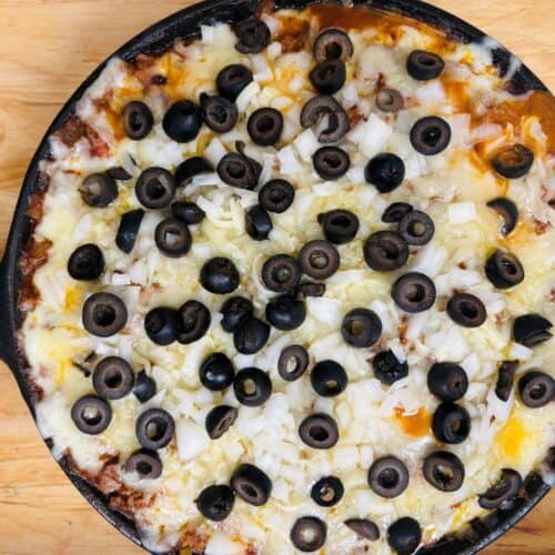 A skillet dip topped with melted cheese, chopped onions, and sliced black olives on a wooden surface.