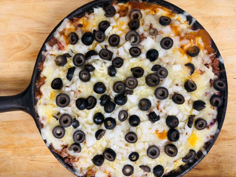 A skillet dip topped with melted cheese, chopped onions, and sliced black olives on a wooden surface.