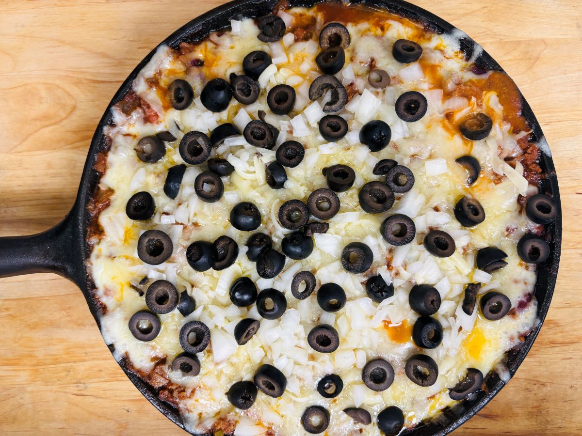 A skillet dip topped with melted cheese, chopped onions, and sliced black olives on a wooden surface.