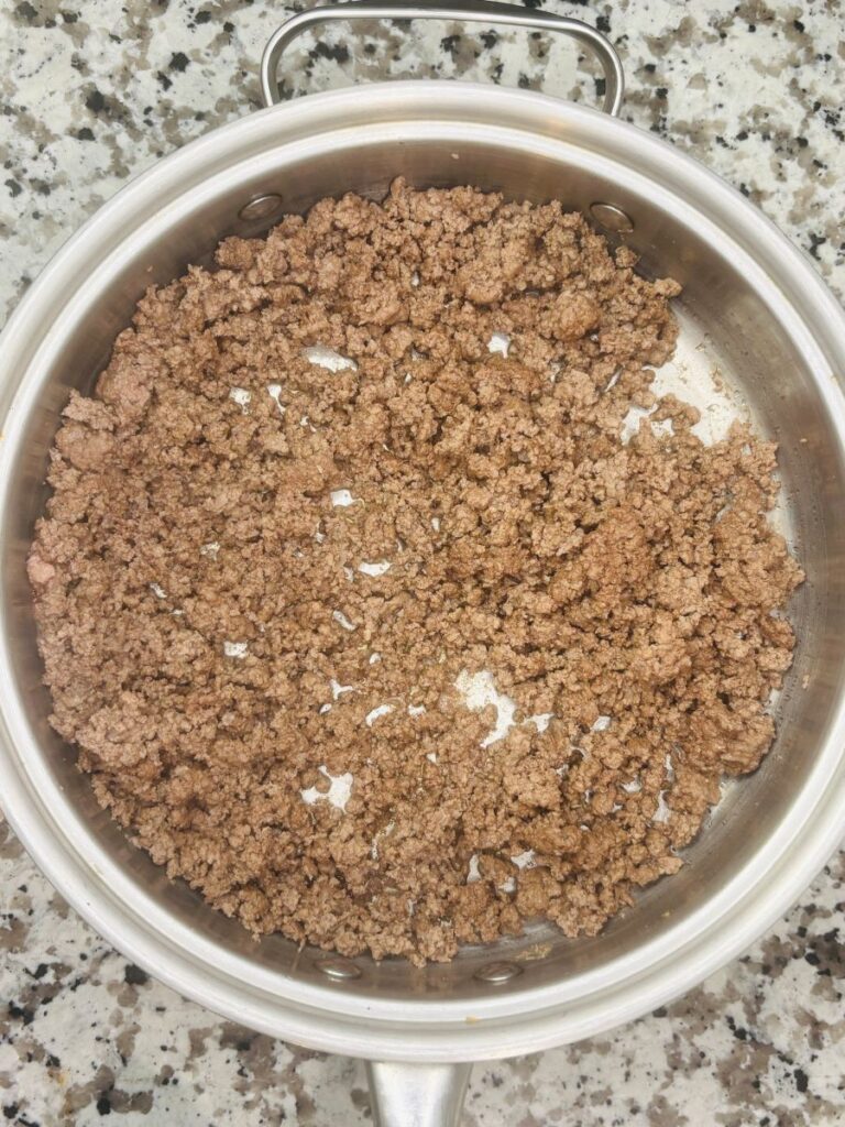 A stainless steel pot filled with cooked ground beef sits on a speckled granite countertop. The beef is browned and crumbled evenly across the surface of the pot.