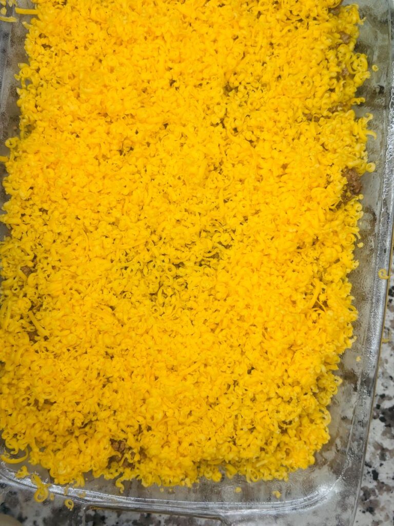A glass baking dish filled with a layer of finely shredded bright yellow cheese covering the top of a casserole, set on a speckled countertop.