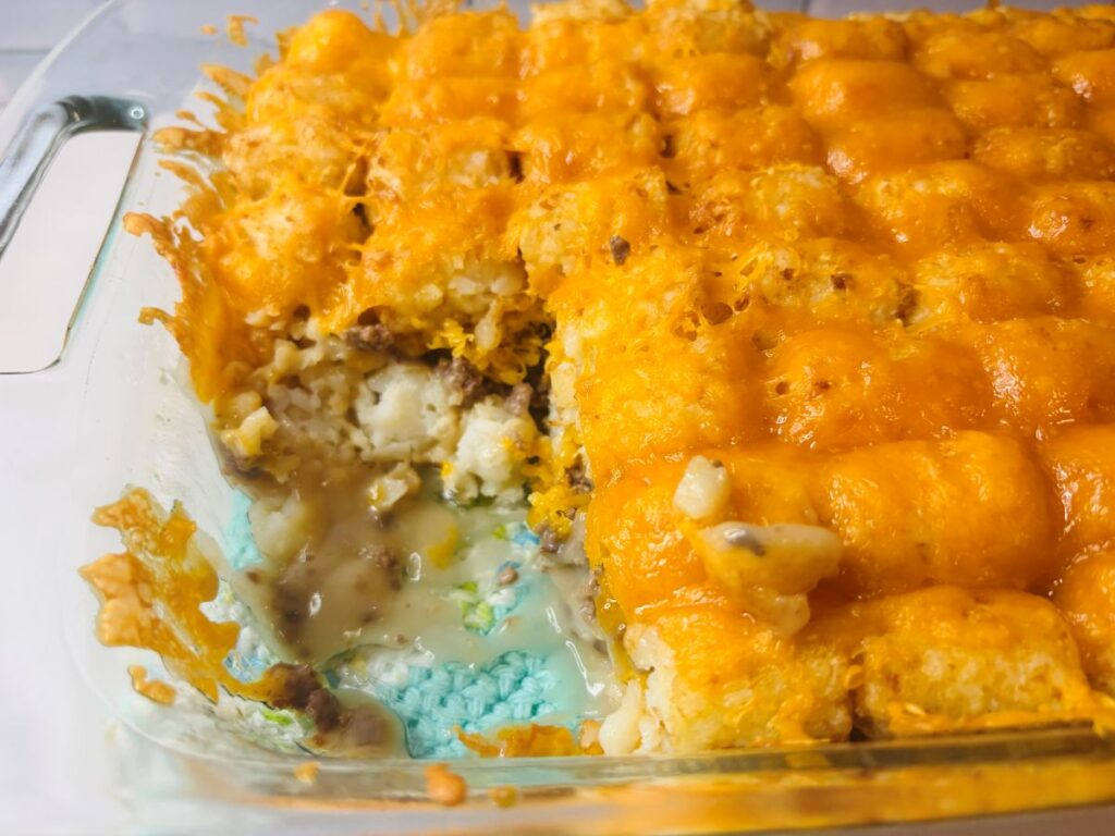 A casserole dish filled with tater tot hotdish, featuring golden brown tater tots and melted cheese on top, with a serving scooped out to reveal a creamy filling with ground meat and vegetables.