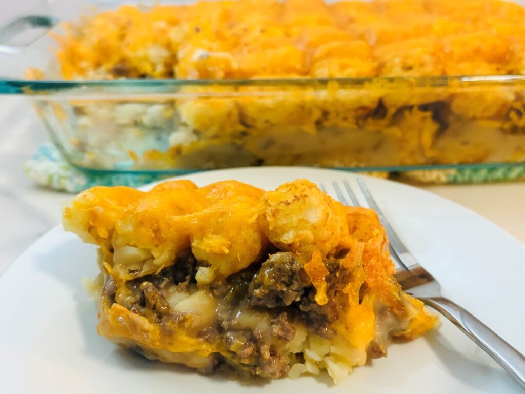 A slice of cheesy tater tot casserole with ground beef and melted cheese on a white plate, with a fork beside it and a glass baking dish of casserole in the background.