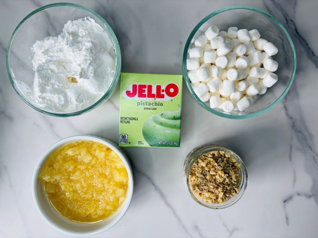 Five ingredients for a recipe on a marble surface: a bowl of whipped topping, a box of pistachio Jell-O pudding mix, a bowl of mini marshmallows, a bowl of crushed pineapple, and a bowl of chopped nuts.
