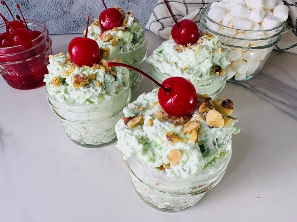 Three glass bowls filled with green pistachio fluff salad, topped with chopped nuts and maraschino cherries. In the background are jars of cherries and mini marshmallows on a white marble surface.