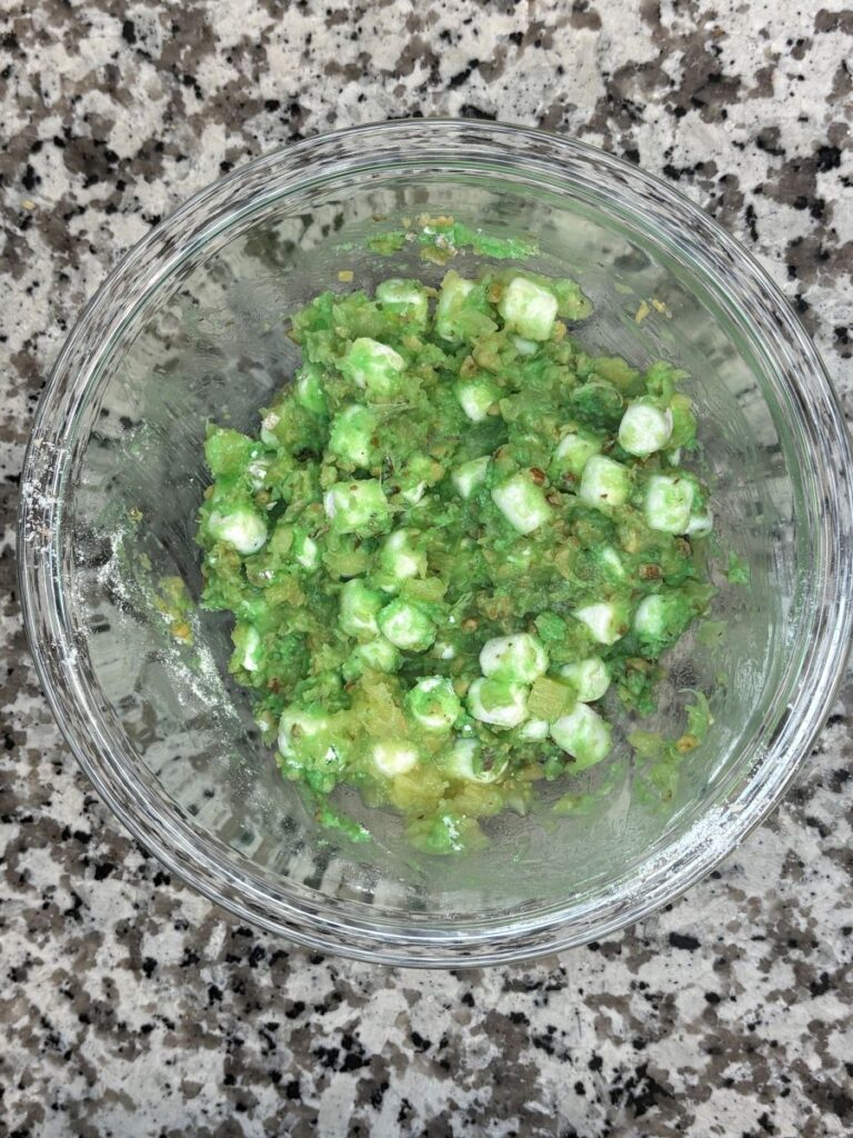 A glass bowl on a granite countertop contains a mixture of mashed green avocado, white cottage cheese curds, and seasonings.
