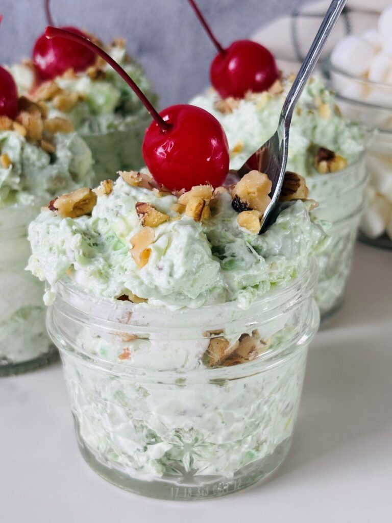 A glass jar filled with green pistachio fluff salad, topped with chopped nuts and a red maraschino cherry, with a spoon inside. Other jars, marshmallows, and cherries are in the background.