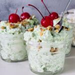 Three glass jars filled with a light green creamy dessert, topped with chopped nuts and maraschino cherries. Mini marshmallows and more cherries are visible in the background. A spoon rests in one of the jars.