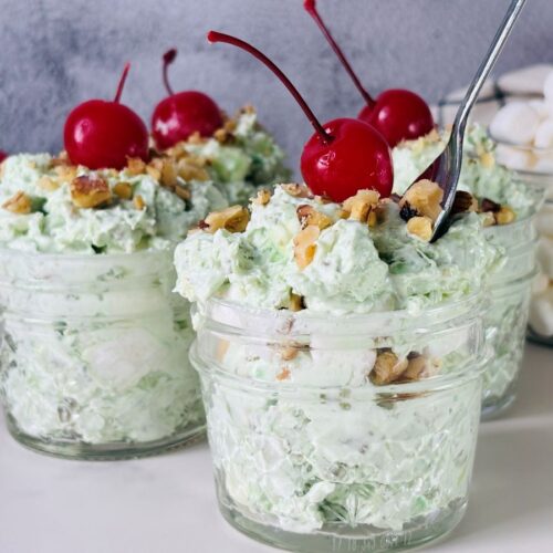 Three glass jars filled with a light green creamy dessert, topped with chopped nuts and maraschino cherries. Mini marshmallows and more cherries are visible in the background. A spoon rests in one of the jars.