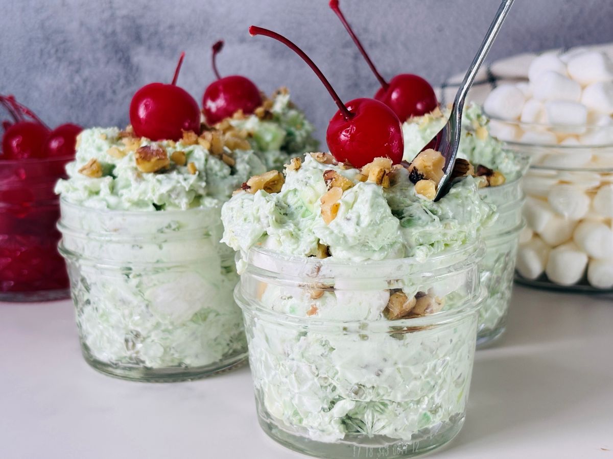 Three glass jars filled with a light green creamy dessert, topped with chopped nuts and maraschino cherries. Mini marshmallows and more cherries are visible in the background. A spoon rests in one of the jars.