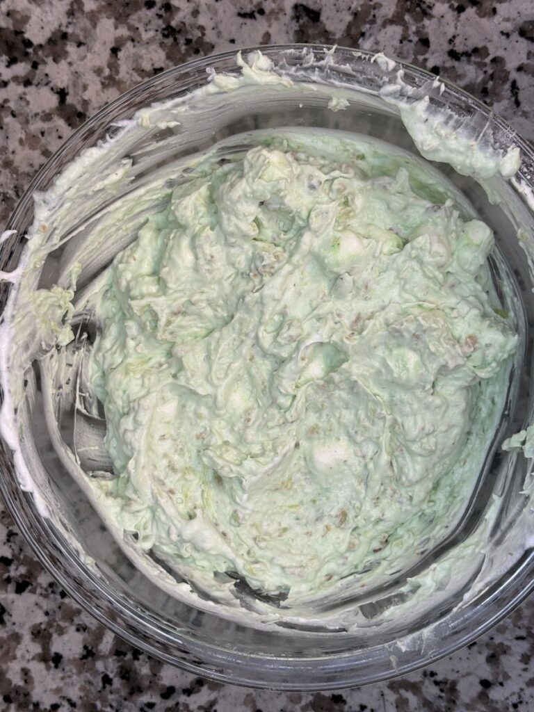 A glass bowl filled with a light green, creamy mixture sits on a speckled gray countertop. The contents appear fluffy and slightly textured, likely a dessert or salad mixture.
